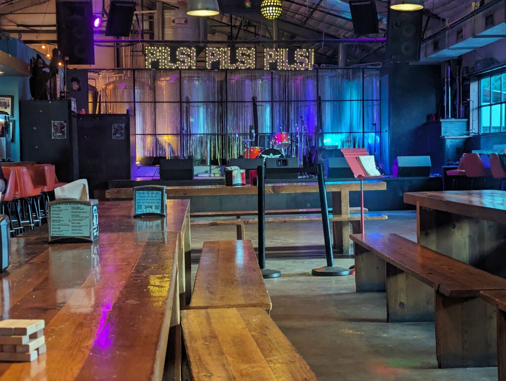 Tables and stage at The Austin Beer Garden Brewery. Over the stage, it says "PILS! PILS! PILS!" in large, illuminated letters.