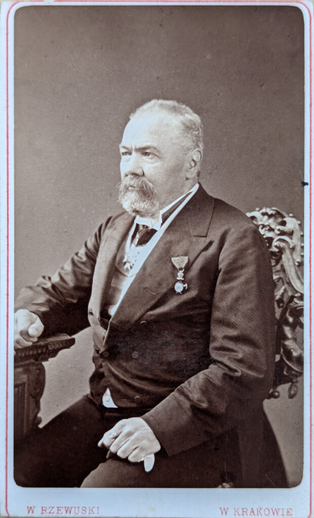 Photo of Johann Götz in a sitting position with his right arm resting on a table and his left hand resting on his left upper thigh, showing him halfway from the side, by Walery Rzewuski. Digitisation licensed under CC BY 4.0. https://creativecommons.org/licenses/by/4.0/