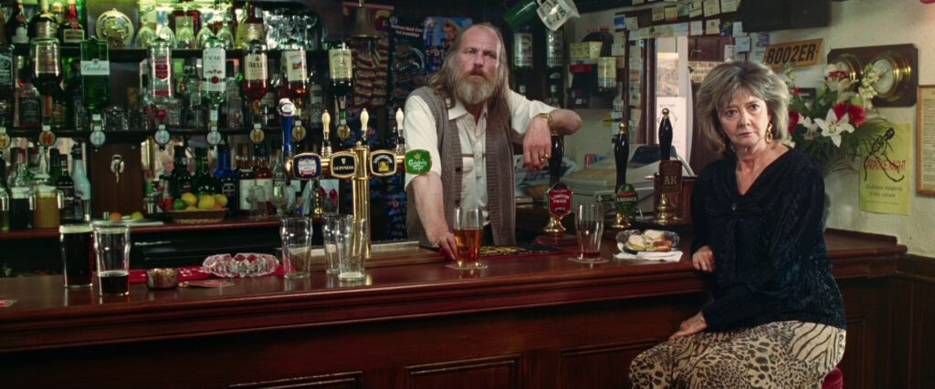 The bar in the 2005 film version, with keg taps of Kronenbourg 1664, Guinness, Strongbow and Carlsberg, and pump clips advertising Fuller's London Pride, Fuller's Chiswick Bitter, and a third beer where only "AK" is really legible (possibly McMullen AK?). One man is stood behind the bar (Albie Woodington), and a woman (Su Elliot) is sat leaning against the bar.