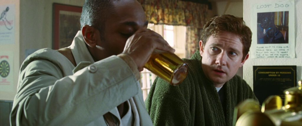 Ford Prefect (Mos Def) drinking a pint of pale beer, with Arthur Dent (Martin Freeman) looking at him.