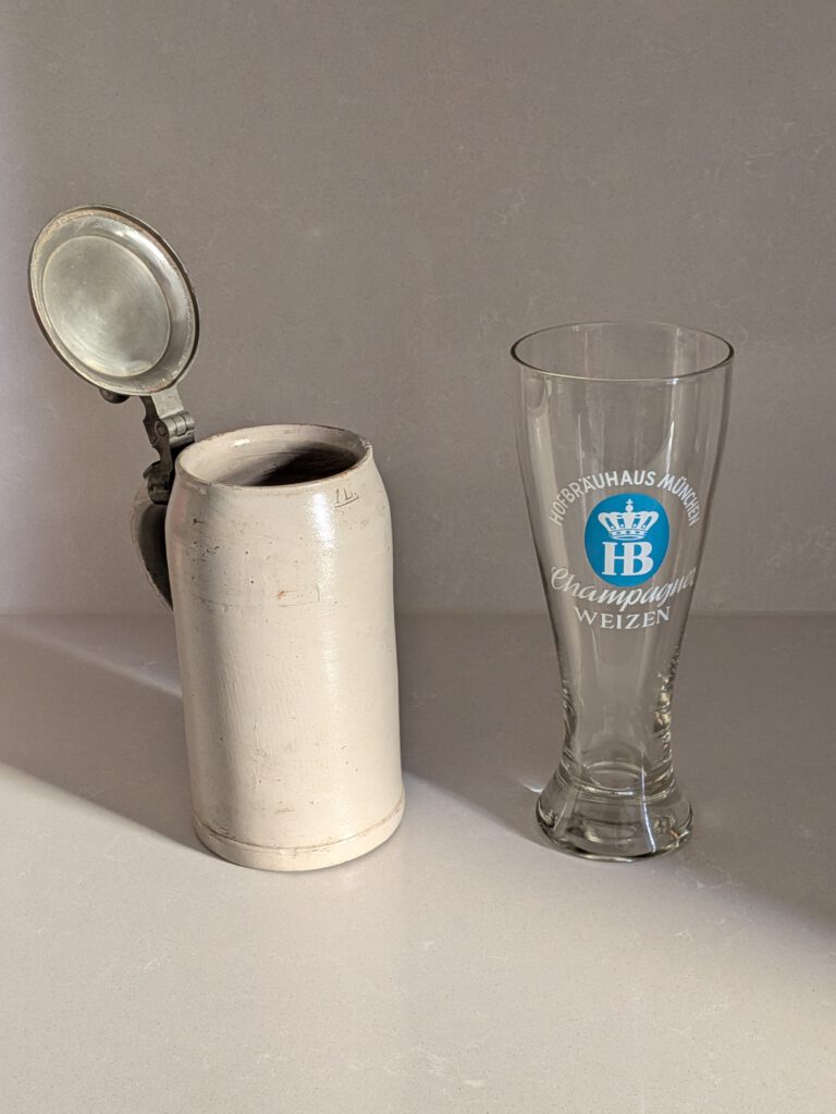 A historic stoneware mug with a tin lid (ca. 1900) and a wheat beer glass (ca. 1960) with Hofbräuhaus München Champagner Weizen branding.