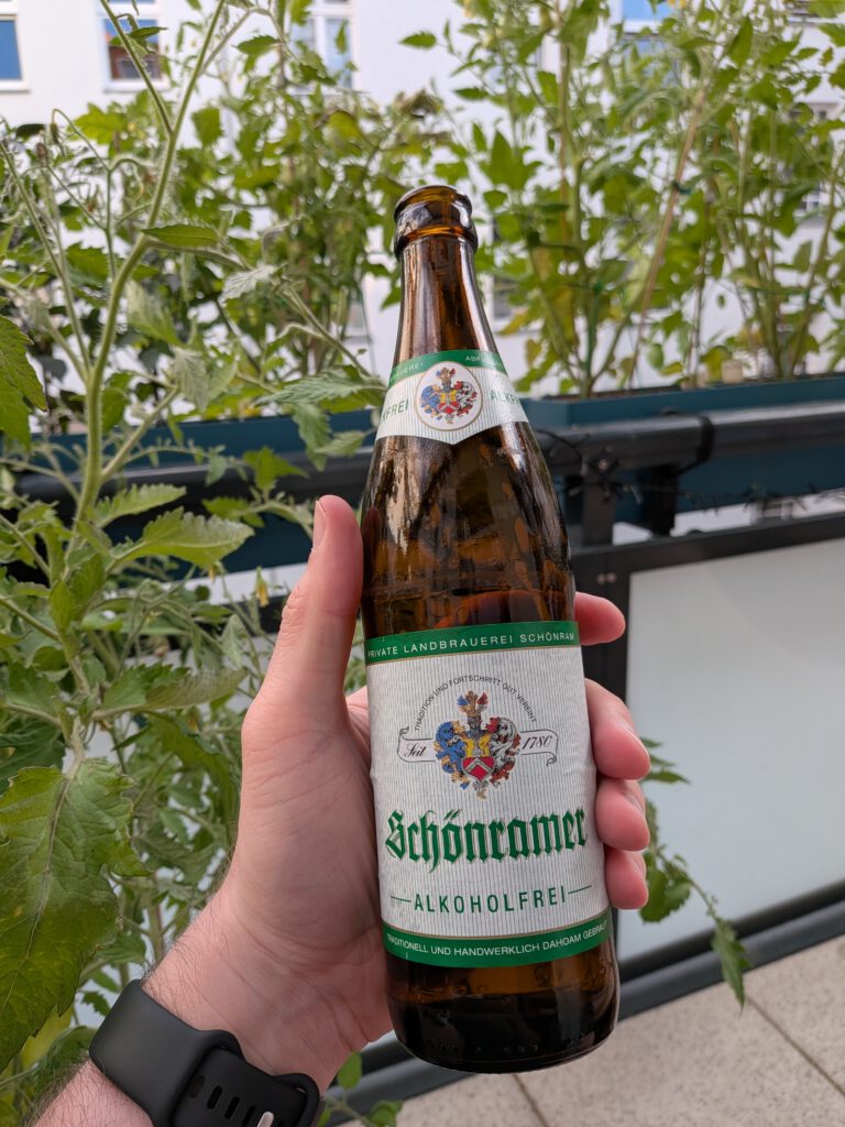 Me holding a bottle of Schönramer Alkoholfrei on my balcony. It's a standard brown German NRW beer bottle. The label says "Private Landbrauerei Schönram", the brewery logo with "seit 1780" referring to the foundation date, and "Schönramer Alkoholfrei" in the center. The label itself is a white with thin vertical green stripes, and green stripes on top and bottom with white text on it. In the background, tomato and chili plants growing on the balcony can be seen.
